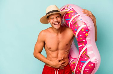 Mavi arka planda izole edilmiş şişme bir çörek tutan genç beyaz adam gülüyor ve eğleniyor..