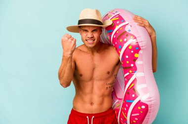 Mavi arka planda izole edilmiş şişme donut tutan genç beyaz bir adam kameraya yumruk gösteriyor, agresif bir yüz ifadesi..