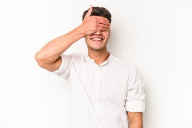 Beyaz arka planda soyutlanmış beyaz tenli genç bir adam elleriyle gözlerini kapatıyor, geniş bir gülümsemeyle bir sürpriz bekliyor..