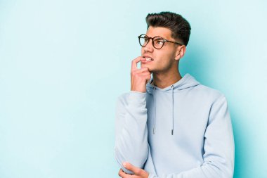 Mavi arka planda izole edilmiş genç beyaz adam şüpheli ve şüpheci bir ifadeyle yana bakıyor..