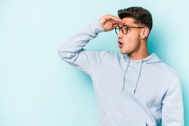 Kafkasyalı genç adam mavi arka planda izole edilmiş, uzaklara bakıyor ve elini alnına koyuyor..