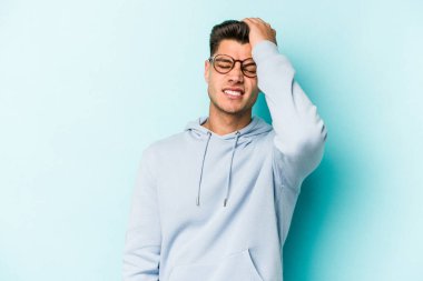 Kafkasyalı genç adam mavi arka planda izole edilmiş bir şekilde bir şeyi unutuyor. Alnına avuç içi ve gözlerini kapayarak vuruyor..