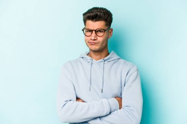 Kafkasyalı genç adam mavi arka planda izole edilmiş hoşnutsuzca kaşlarını çatmış, kollarını kavuşturmuş..