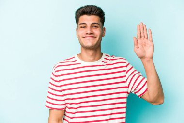 Mavi arka planda izole edilmiş genç beyaz adam gülümseyerek beş numarayı parmaklarıyla gösteriyor..