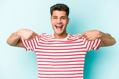 Mavi arka planda izole edilmiş genç beyaz adam parmağını göstererek şaşırdı, geniş bir gülümsemeyle..