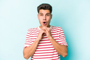 Kafkasyalı genç adam mavi arka planda izole edilmiş şans için dua ediyor, şaşırmış ve ağzını açıp önüne bakıyor..