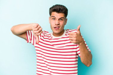 Kafkasyalı genç adam mavi arka planda izole edilmiş, baş parmağını kaldırıyor, zor seçim konsepti.