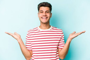 Mavi arka planda izole genç beyaz adam kollarıyla ölçek yapar, mutlu ve kendinden emin hisseder..