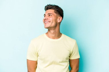Kafkasyalı genç adam mavi arka planda izole edilmiş rahat ve mutlu bir şekilde gülüyor, boynunda dişlerini gösteriyor..