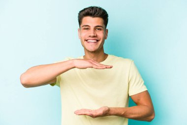 Mavi arka planda izole genç beyaz adam iki eliyle bir şey tutuyor, ürün sunumu..