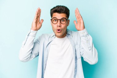 Mavi arka planda izole edilmiş genç beyaz bir adam zaferi ya da başarıyı kutluyor, şaşırıyor ve şok oluyor..