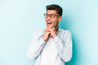 Mavi arka planda izole edilmiş genç beyaz adam ellerini çenesinin altında tutuyor, mutlu bir şekilde yana bakıyor..