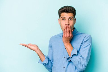 Mavi arka planda izole edilmiş genç beyaz adam, avuç içi fotokopi alanını elinde tutmaktan etkilenmiş..