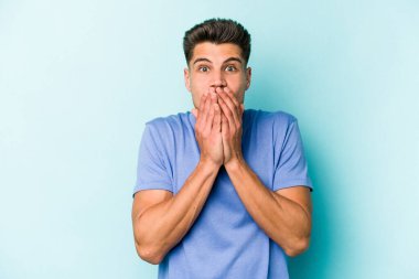 Mavi arka planda izole edilmiş genç beyaz adam elleriyle ağzını kapatırken şok oldu..