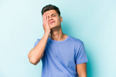 Kafkasyalı genç adam mavi arka planda izole edilmiş. Yorgun ve uykulu. Elini başının üstünde tutuyor..