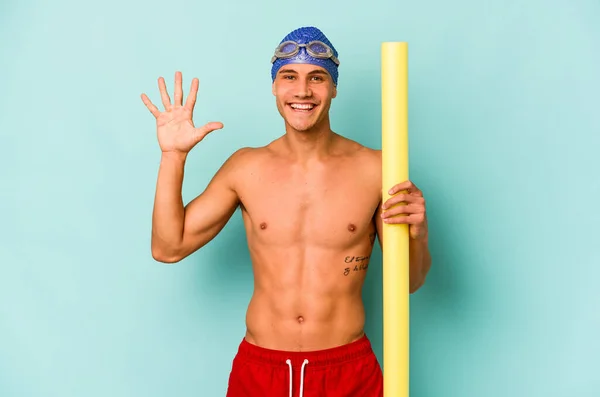 年轻的高加索游泳运动员 手握发泡棒 被蓝色背景隔离 笑容满面 用手指露出第五名 — 图库照片