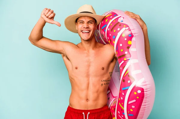 Mavi arka planda izole edilmiş şişme bir çörek tutan genç beyaz adam gururlu ve kendinden emin hissediyor, örnek olarak.