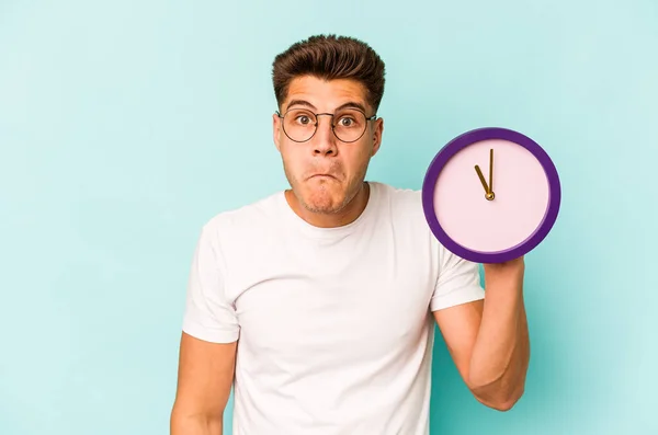 Young Caucasian Man Holding Clock Isolated Blue Background Shrugs Shoulders — стоковое фото