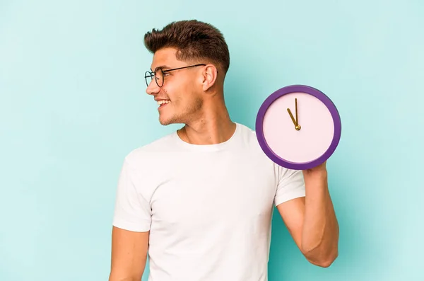 Young Caucasian Man Holding Clock Isolated Blue Background Looks Aside — ストック写真