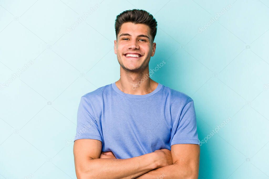 Young caucasian man isolated on blue background who feels confident, crossing arms with determination.