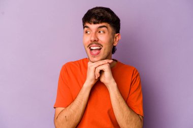 Mor arka planda izole edilmiş genç beyaz adam ellerini çenesinin altında tutuyor, mutlu bir şekilde yana bakıyor..