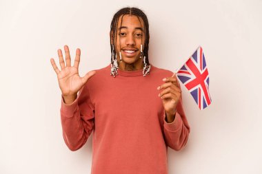 Beyaz arka planda izole edilmiş İngiliz bayrağı tutan genç Afrikalı Amerikalı adam gülümseyerek beş numarayı parmaklarıyla gösteriyor..