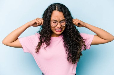 Genç İspanyol kadın mavi arka planda izole edilmiş parmaklarıyla kulaklarını kapatıyor, gürültülü bir ortam yüzünden stresli ve çaresiz..