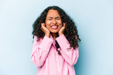 Mavi arka planda izole edilmiş genç İspanyol kadın kulaklarını elleriyle kapatıyor..