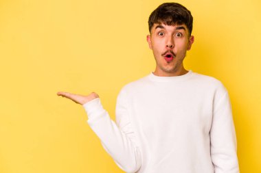 Sarı arka planda izole edilmiş genç beyaz adam, avuç içi fotokopi alanını elinde tutmaktan etkilendi..