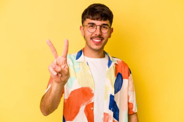 Sarı arka planda izole edilmiş genç beyaz adam zafer işareti gösteriyor ve gülümsüyor..