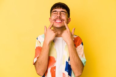 Sarı arka planda izole edilmiş genç beyaz adam gülümsüyor, parmaklarını ağzına doğrultuyor..