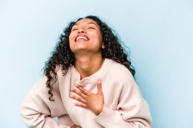 Mavi arka planda izole edilmiş genç İspanyol kadın gülüyor, ellerini kalbe koyuyor, mutluluk kavramını koruyor..