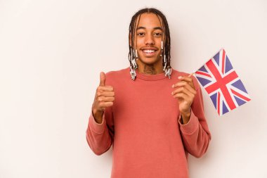 Beyaz arka planda izole edilmiş İngiliz bayrağı tutan genç Afrikalı Amerikalı adam gülümsüyor ve başparmağını kaldırıyor.