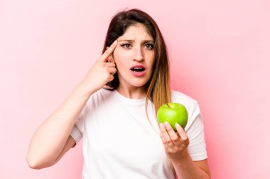 Pembe arka planda elma tutan beyaz bir kadın işaret parmağıyla yaptığı hayal kırıklığı jestini gösteriyor..