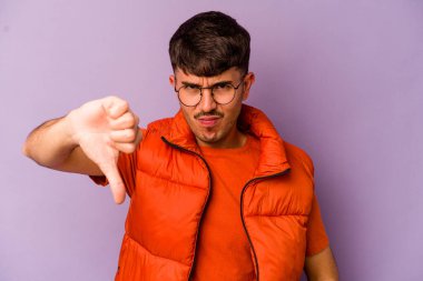 Mor arka planda izole edilmiş genç beyaz adam baş parmağını aşağı gösteriyor ve hoşlanmadığını ifade ediyor..