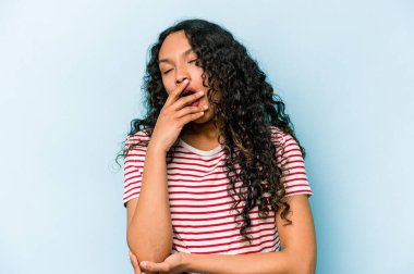 Mavi arka planda izole edilmiş genç İspanyol kadın esniyor. Ağzı eliyle kapatan yorgun bir hareket sergiliyor..