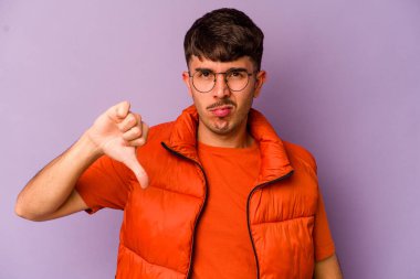 Mor arka planda izole genç beyaz adam baş parmağını aşağı gösteriyor, hayal kırıklığı konsepti.