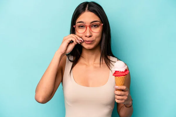 Young hispanic woman holding an ice cream isolated on blue background with fingers on lips keeping a secret.