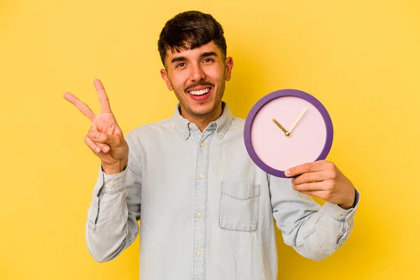 黄色の背景に時計を持つ若いヒスパニック系の男喜びと屈託のない指で平和のシンボルを示す — ストック写真