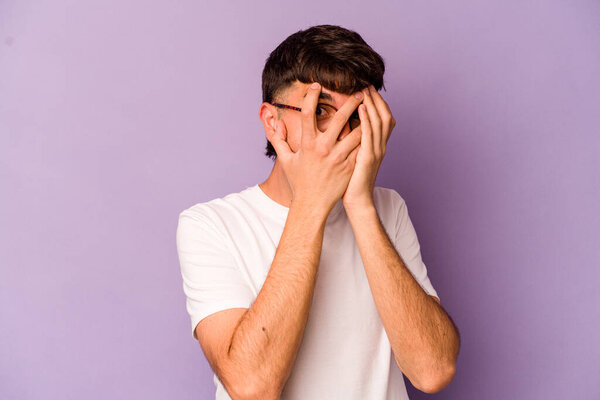 Young caucasian man isolated on purple background blink through fingers frightened and nervous.