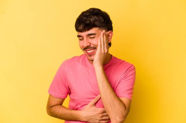 Sarı arka planda izole edilmiş genç beyaz adam mutlu bir şekilde gülüyor ve ellerini karnının üstünde tutarak eğleniyor..