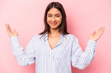 Pembe arka planda izole edilmiş genç İspanyol kadın işaret parmaklarıyla küçük bir şey tutuyor, gülümsüyor ve kendinden emin..