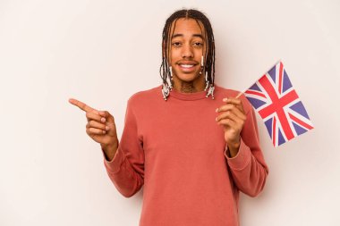 Beyaz arka planda izole edilmiş İngiliz bayrağı tutan genç Afrikalı Amerikalı bir adam gülümsüyor ve boş alanda bir şey gösteriyor..