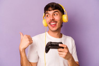 Latin asıllı genç bir adam mor arka plan noktalarında izole edilmiş bir bilgisayar oyunu kontrolörüyle oynuyor. Başparmak uzakta, gülüyor ve kaygısız..