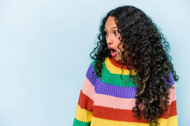 Genç İspanyol kadın mavi arka planda izole edilmiş. Gördüğü bir şey yüzünden şoka uğruyor..