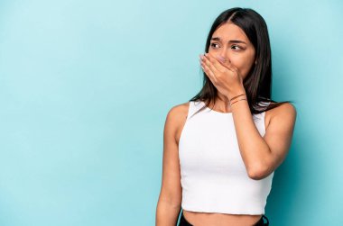 Genç İspanyol kadın mavi arka planda izole edilmiş. Düşünceli bir şekilde bir fotokopi odasına bakıyor. Ağzını eliyle kapatıyor..