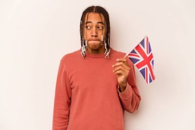 Beyaz arka planda izole edilmiş İngiliz bayrağı tutan genç Afrikalı Amerikalı adam kafası karışmış ve kendinden emin değil..