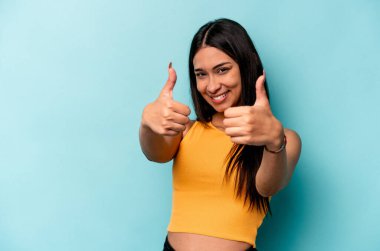 Genç İspanyol kadın mavi arka planda izole edilmiş. Baş parmağını kaldırıyor, gülümsüyor ve kendinden emin..
