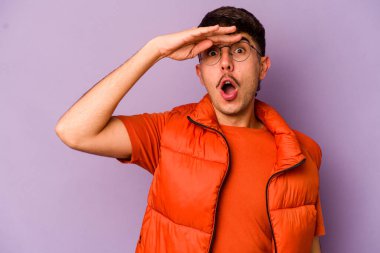 Mor arka planda izole edilmiş genç beyaz adam uzaklara bakıyor ve elini alnına koyuyor..