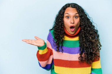 Mavi arka planda izole edilmiş genç İspanyol kadın avuç içi fotokopi alanını tutmaktan etkilenmiş..